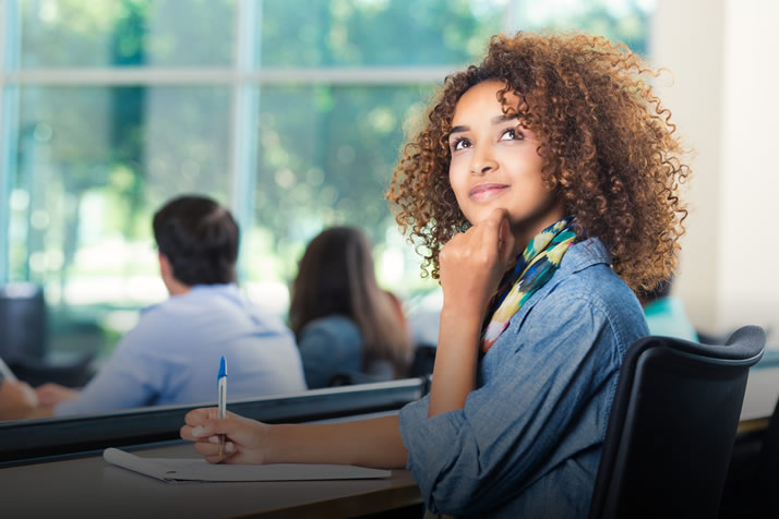 Como escolher sua profissão? Descrição da imagem: mulher jovem na sala de aula, olhando para cima pensativa enquanto faz a prova do Enem.