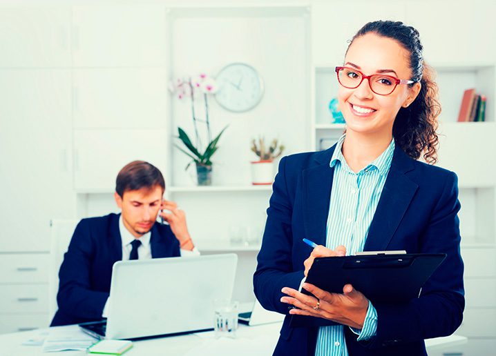 Imagem de uma profissional recém-contratada em um escritório fazendo anotações. Ao fundo, jovem sentado à mesa, utilizando o laptop e falando no celular.